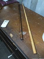 Two sticks lying on a wooden surface; left is a darker brown leather wrapped military staff about 24 inches long, right is a lighter wooden Carnie stick about 31 inches long.