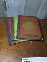 Stack of three Minecraft hardcover books on wooden surface with visible corner wear and pixel art covers.