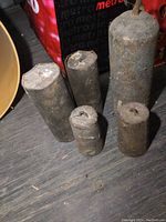 Five old cast iron weights displayed on a wooden floor next to a red bag, showing varying sizes and surfaces heavily rusted and aged.