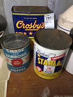 Photo of Crosby's molasses tin (blue with yellow/red text), Borden's Instant Starlac tin (white/yellow with graphics), and blue Chase & Sanborn coffee tin on wooden surface.