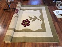 Partially rolled rectangular floral area rug with cream background and olive green border on wood floor.