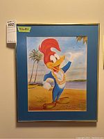 Framed colorful art print of Woody Woodpecker wearing a lei on a beach with palm trees and ocean in the background. Blue mat and gold frame are visible. Yellow sticky note with dimensions 15.5 x 18.5 inches attached to frame.
