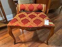 Front view of the wood framed bench showing red and gold patterned upholstery with tears and the carved wood ornamentation on front apron.