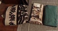 Three folded woven throw blankets on carpeted floor: one with cream base and brown/black pattern, one brown/cream with fringe, and one solid green.