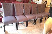 Front view of all six matching gray upholstered dining chairs with tufted backrests and black wooden legs arranged in a row on tile floor.