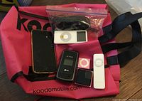 Photo showing various devices placed on a pink bag including a black iPhone, small iPods (white and pink), and cables in a plastic bag.