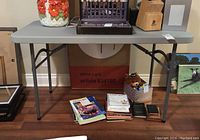 Front view of grey Staples folding table showing top surface and legs, placed indoors on wooden floor.