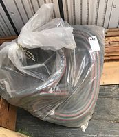 Coiled black garden hose with red stripes wrapped in a plastic bag placed on wooden pallet inside metal shed.