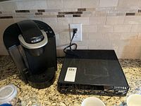 Black Keurig coffee maker sitting on kitchen counter near a power outlet, with water reservoir visible on the left side, and black metal pod holder rack next to it.