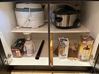View inside a cabinet showing multiple kitchen appliances and gadgets including Nesco dehydrator, Crock-Pot slow cooker, coffee grinder, pineapple cutter, and milk frother boxed items.