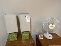 Pair of green ceramic fabric shade table lamps next to Airworks fan and ThermPro indoor barometer on wooden surfaces against a cream wall