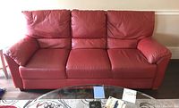 Front view of red leather three-seat sofa with visible wear and creasing on cushions and armrests, placed in a living room setting with table and carpet visible.