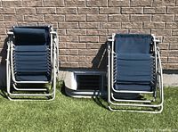 Two folded outdoor zero gravity lounge chairs leaning against a brick wall on grass.