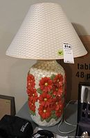Full lamp with woven basket design base decorated with red flowers and textured white lampshade.