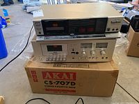 Aiwa AD-F330C cassette deck stacked on top of Akai CS-707D deck, both on an Akai box in a cluttered garage environment.