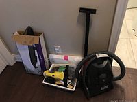 Photo showing vacuum cleaner hose and part of the Rival electric griddle box along with Karcher steam cleaner attachments in a tub on the floor against a wall.