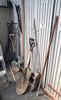 Wide view showing multiple yard tools including shovels, rakes, broom leaning against metal shed wall