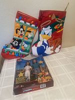 Photo showing two Christmas stockings: one red with Donald Duck with gold trim and one multicolored with Goofy, Mickey, and Donald in 3D window scene, along with boxed Troy and Gabriella dolls from High School Musical 2