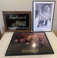 Three pieces of winery-themed wall art displayed on a table: an Inglenook glass mirror with wood frame, a framed whimsical cartoon print, and a framed Arrowood Vineyards photographic poster with signatures.