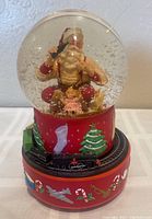Front view of the Coca-Cola Santa Claus musical snow globe showing seated Santa holding a Coke bottle, surrounded by snow inside a glass globe. The red base decorated with Christmas symbols and a miniature black train on a track around the base is visible.