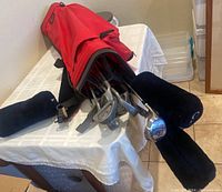 Red and black golf bag lying on table with golf clubs visible inside it along with headcovers on some clubs.
