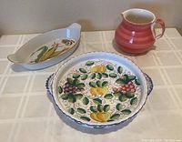 Overall view of three ceramic serving pieces: reddish striped pitcher, fruit decorated large round dish with handles, corn-shaped porcelain dish.