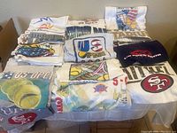 View of all 14 sports t-shirts folded and stacked with visible logos showing various sports events including U.S. Open tennis, baseball Subway Series, Olympics, and 49ers.