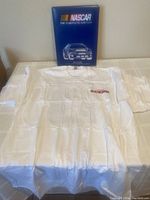 Full view of the white NASCAR Scene t-shirt laid flat with the blue covered NASCAR: The Complete History book behind it, showing size and condition.