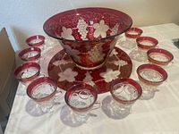 Front angle showing punch bowl, plate, and 12 cups arranged around on table, illustrating ruby banding and grapevine etching.