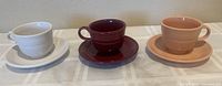 Three Fiesta Ware cups and saucers displayed from left to right: white, deep burgundy, and peach. All items are ceramic and have a ribbed texture with handles on the cups.