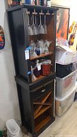 Full view of black bar tower cabinet showing upper glass rack, two shelves with glassware, drawer, and lower wine rack.