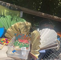 Photo showing seven assorted hand fans placed in and around a clear plastic bin. Includes colorful and plain fans with various designs and materials.