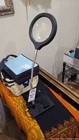 Full view of electric magnifying lamp standing on table with black base, flexible silver neck, and circular magnifying lens with black trim.