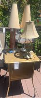 Photo showing metal rolling corner table with three vintage lamps placed on top, outdoor setting.