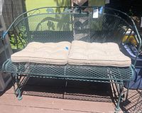 Green metal settee glider with two beige cushions on seat area, outdoors in daylight.