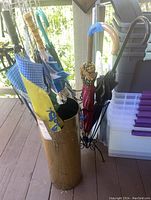 Photo shows assortment of umbrellas including a yellow and blue checkered umbrella, a clear umbrella with blue handle, and others stored inside a brass cylindrical umbrella stand and a black wrought iron stand.