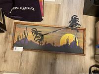 Frontal view of the wall art, showing the sunset desert landscape painting with silhouetted cacti and mountains in a wooden frame, with the black metal Kokopelli towel rack mounted above.
