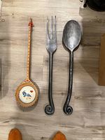View of three large wooden wall hanging utensils: one natural wood with painted bird spoon, one metallic silver fork, and one metallic silver spoon with curled handles.
