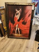 Front view of original framed wall art painting of a ballerina in a flowing red dress with arms raised, standing en pointe on wooden floor.