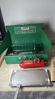 Green Coleman two-burner stove with windshields upright, red fuel tank and metal hotplate grill placed in front.