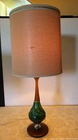 Full front view of the mid century table lamp showing the fabric shade, wood and green ceramic base with round wooden platform.