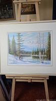 Framed oil painting showing winter mountain landscape with snow, lake and trees.