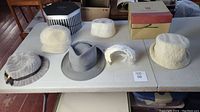Six hats on table; includes one men's gray fedora and five ladies hats in cream and white shades of various textures and forms.