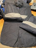Two fabric garment bags shown on a table, one open revealing interior and one folded closed with a gray stripe across each.