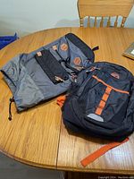 Two backpacks on a wooden table, one large gray backpack with black straps and tan accents, and one medium black backpack with orange trim and vertical orange stripe on front.