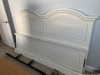 White painted wooden king size headboard with side rails and foot frame resting on floor, showing decorative carved details and some scratches.