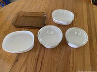 All five baking dishes displayed on a wooden table: three white ceramic dishes with glass lids, one white ceramic dish without a lid, and one rectangular brown glass dish.