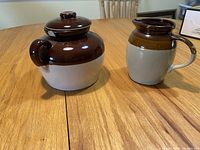 Photo showing the ceramic crock pot with its lid on and the matching pitcher side by side on a wooden table, both with two-tone brown and beige glaze.