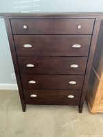 Front view of the dark wood tall dresser with five drawers and metal knobs/handles on a carpeted floor.