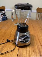 Front view of Oster electric mixer with glass blender jar, controls and power cord visible on wooden table.